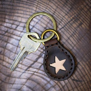 May include: A brown leather keychain with a white star in the center. The keychain is attached to a gold key ring and a silver key.