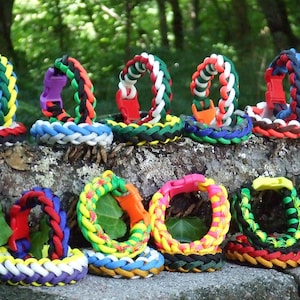 May include: A collection of colorful paracord bracelets with various patterns and buckle closures. The bracelets are arranged in two rows on a tree trunk.