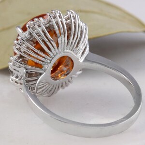 May include: A silver ring featuring a large orange gemstone encircled by smaller clear gemstones. The ring is set in a halo style.