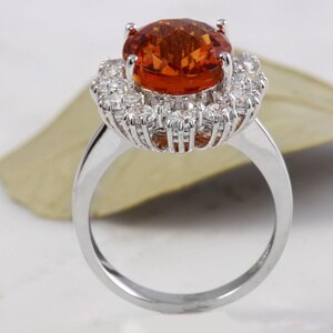 May include: A silver ring featuring a large orange gemstone encircled by a halo of smaller white gemstones.