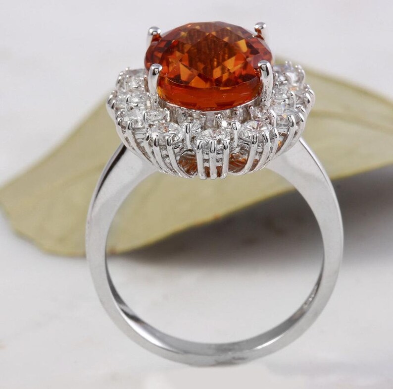 May include: A silver ring featuring a large orange gemstone encircled by a halo of smaller white gemstones.