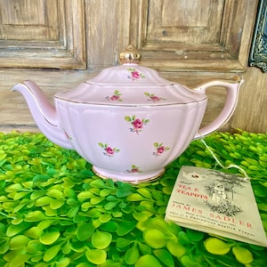 Könnte beinhalten: Eine rosafarbene Teekanne mit goldener Verzierung und einem Blumenmuster. Die Teekanne steht auf einer grünen Oberfläche mit einem Etikett, auf dem steht "About Tea & Teapots Issued by James Sadler Makers of Fine Teawares since 1892 This is a Guaranteed English Teapot".