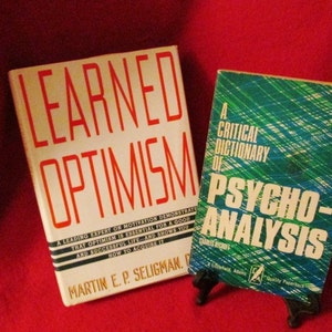 May include: Two books on a red surface. The book on the left is titled "Learned Optimism" by Martin E.P. Seligman. The book on the right is titled "A Critical Dictionary of Psycho-Analysis" by Charles Rycroft.
