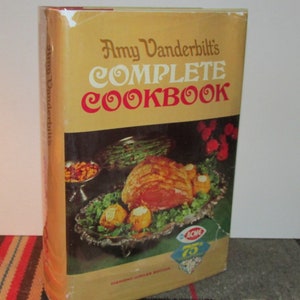 May include: A yellow and red hardcover cookbook titled "Amy Vanderbilt's Complete Cookbook". The cover features a photo of a roasted ham with a side of green beans and a bowl of nuts.