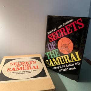 May include: Two books titled "Secrets of the Samurai: A Survey of the Martial Arts of Feudal Japan" by Oscar Ratti and Adele Westbrook. One book is in a cardboard box, the other is a hardcover book with a black cover and colorful text.
