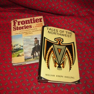 May include: Two paperback books, one titled "Frontier Stories" by Louis L'Amour and the other titled "Tales of the Northwest" by William Joseph Snelling. The cover of the first book features a cowboy riding a horse in front of a mountain range. The cover of the second book features a stylized eagle with a black and gold design.