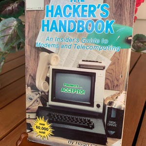 May include: The Hacker's Handbook book cover. The title is in large blue letters. The cover features a vintage computer with the text "Password... ACCEPTED!" on the screen. The book is propped up on a wooden stand. The book is an insider's guide to modems and telecomputing.