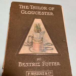 May include: A vintage copy of "The Tailor of Gloucester" by Beatrix Potter. The book cover is brown with white text and a triangular illustration featuring sewing tools. The publisher's name, F. Warne & Co., is at the bottom.