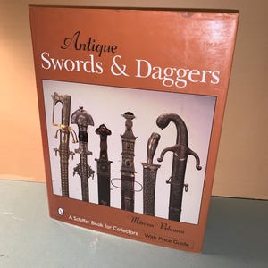 May include: A book titled "Antique Swords & Daggers" with a collection of antique swords and daggers. The cover is orange with white text. The book is a Schiffer Book for Collectors with a price guide.