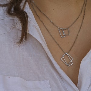May include: A silver necklace with two geometric pendant charms. The larger charm is a diamond shape, and the smaller charm is a rectangle. The necklace is layered on a white shirt.