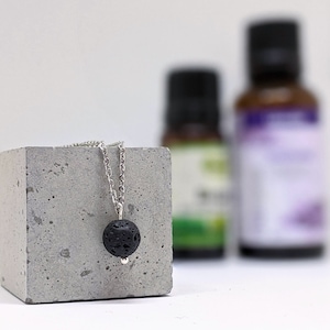 Puede incluir: Un collar de cadena plateada con un colgante de piedra de lava negra. El collar se muestra en un bloque de hormigón gris.