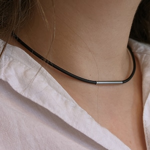 Puede incluir: Un collar negro simple de tipo choker con un colgante de barra de plata.