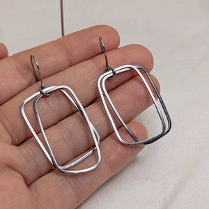 May include: Silver double-loop rectangle earrings. The earrings are made of two silver rectangles that are connected at the top and bottom. The earrings are hanging from silver hooks.