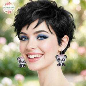 Può includere: Un paio di orecchini pendenti con un design a forma di panda realizzato con perline. Ogni panda è bianco e nero con un piccolo fiocco rosa sulla testa. Gli orecchini sono composti da piccole perline disposte a griglia, creando un effetto pixelato.