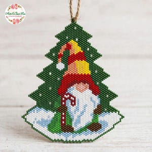 Può includere: Una decorazione per albero di Natale in perline raffigurante uno gnomo con una lunga barba bianca, che indossa un cappello rosso e giallo e tiene in mano un bastoncino di zucchero rosso e bianco su uno sfondo di albero verde con neve bianca.