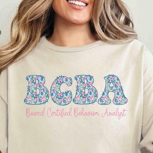 May include: A beige t-shirt with the text "BCBA Board Certified Behavior Analyst" in a floral design. The shirt is a short-sleeved, casual style.