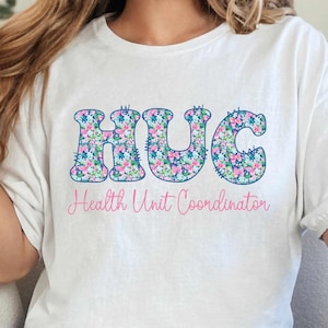 May include: White t-shirt with the word "HUG" in a floral pattern, above the text "Health Unit Coordinator" in pink. The shirt is paired with blue jeans. The overall style is casual and comfortable.