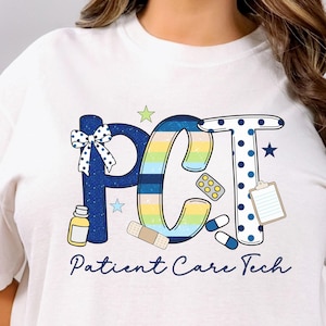 Puede incluir: Camiseta blanca con un diseño gráfico colorido. El diseño deletrea "PCT" en letras grandes, con ilustraciones de temática médica. Debajo de las letras, las palabras "Patient Care Tech" están escritas en una fuente de guion.