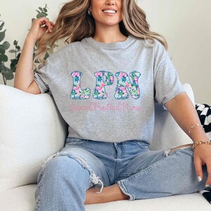 Puede incluir: Camiseta gris jaspeado con las letras "LPN" en un estampado floral. Debajo, las palabras "Licensed Practical Nurse" est&aacute;n impresas en rosa. La camiseta es de cuello redondo.