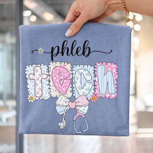 Peut inclure: T-shirt bleu clair avec l'inscription "phleb tech" dans une police décorative. Les lettres sont décorées de motifs floraux et rayés. Un nœud et un stéthoscope sont en dessous.