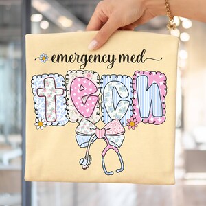 May include: A light yellow t-shirt with the words "emergency med TECH" in a decorative font. The word "TECH" is in a block letter style with floral and striped patterns. A bow and stethoscope graphic are at the bottom.