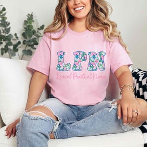 Puede incluir: Una camiseta rosa claro con las letras "LPN" en un estampado floral. Debajo de las letras, el texto dice "Licensed Practical Nurse" en una fuente cursiva. La camiseta se usa con vaqueros azules.