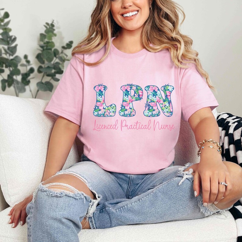 Puede incluir: Una camiseta rosa claro con las letras "LPN" en un estampado floral. Debajo de las letras, el texto dice "Licensed Practical Nurse" en una fuente cursiva. La camiseta se usa con vaqueros azules.