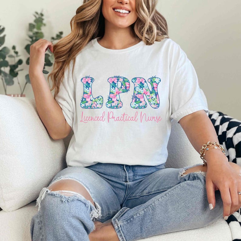 Puede incluir: Camiseta blanca con las letras "LPN" en un dise&ntilde;o floral, sobre las palabras "Licensed Practical Nurse" en rosa. La camiseta se usa con vaqueros azules.