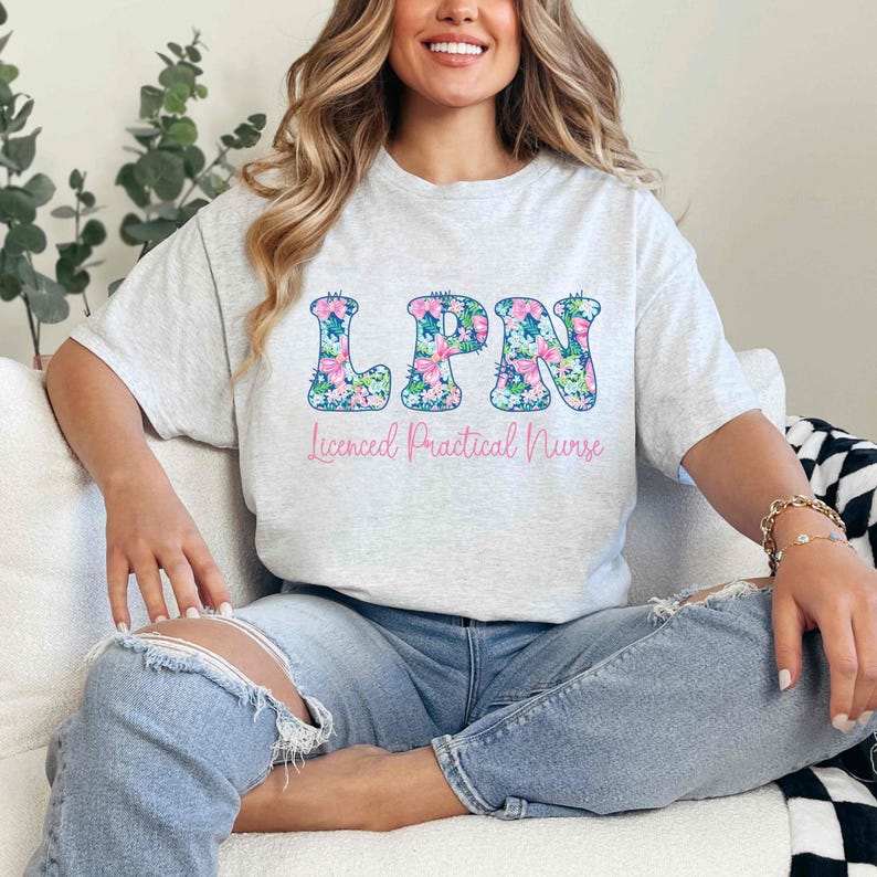 Puede incluir: Una camiseta gris claro con las letras "LPN" en un dise&ntilde;o floral. Debajo, el texto "Licensed Practical Nurse" est&aacute; escrito en rosa. La camiseta se combina con unos vaqueros azules.