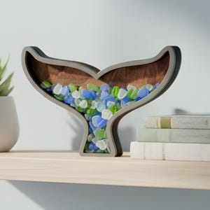 May include: A wooden whale tail-shaped decorative piece filled with blue, green, and clear glass pebbles. The frame is gray with a brown interior, and the piece sits on a light-colored shelf. A succulent plant in a white pot is to the left.
