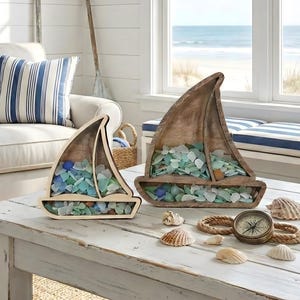 May include: Two wooden sailboat-shaped decorative boxes filled with sea glass in shades of blue, green, and clear. The boxes sit on a white distressed table with seashells, a compass, and rope. A coastal theme.
