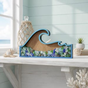 May include: A wave-shaped wooden frame with a blue border, filled with a collection of small, smooth sea glass pieces in shades of blue, green, and white. The interior of the frame features a plain brown backing behind the glass fragments.