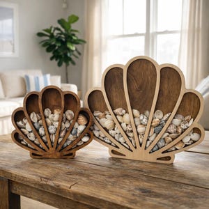 May include: Two wooden seashell-shaped decorative bowls filled with small seashells. The bowls are different sizes and sit on a wooden table. The larger bowl is light-colored wood, and the smaller bowl is a darker wood.