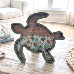 May include: A wooden turtle-shaped decorative piece with a dark green frame. The interior is filled with sea glass in various colors, including white, blue, and orange. The turtle is displayed on a wooden table, suggesting a coastal or nautical theme.