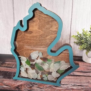 May include: A wooden cutout of the state of Michigan with a teal blue border. The cutout is filled with sea glass of various colors and sizes.