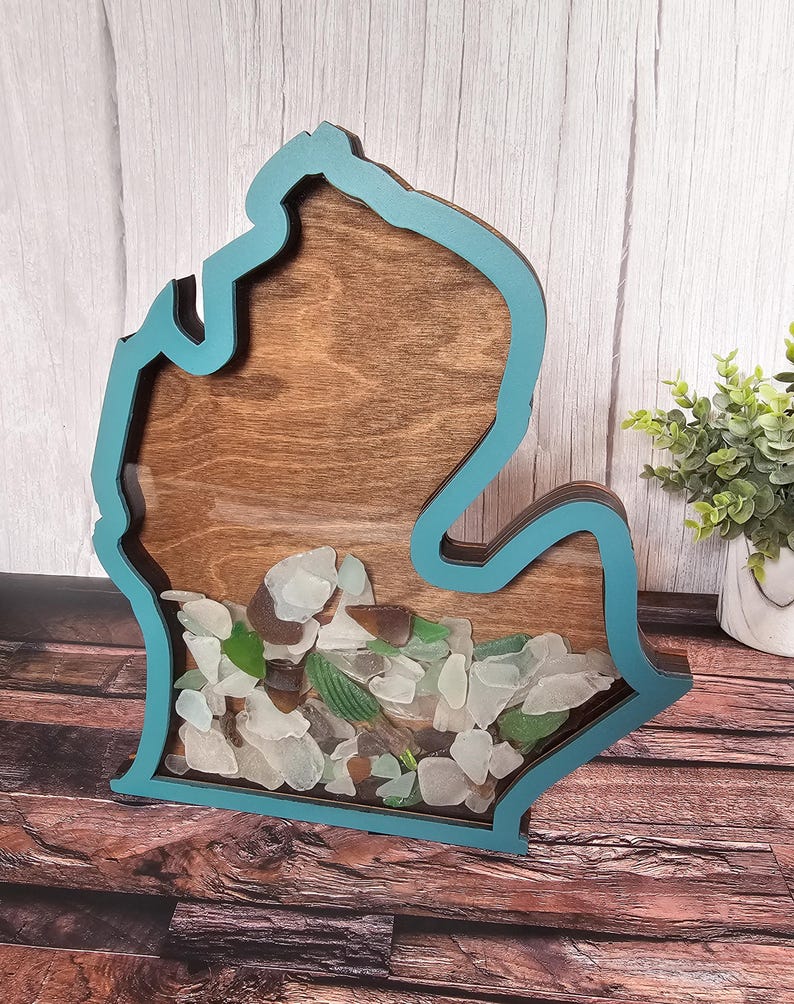 May include: A wooden cutout of the state of Michigan with a teal blue border. The cutout is filled with sea glass of various colors and sizes.