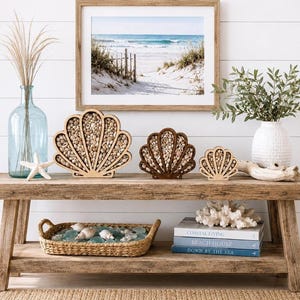 May include: Three wooden shell-shaped shadow boxes filled with small seashells, a light blue glass vase with dried grass, a white textured vase with greenery, a woven tray with sea glass, and a stack of three books titled Coastal Living.