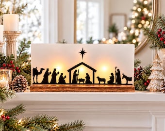 May include: A rectangular white light-up nativity scene display featuring a black silhouette of the holy family, shepherds, animals, and a star. The scene is mounted on a rectangular wooden base and glows with warm internal illumination.