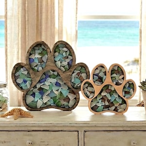 May include: Two wooden paw print decorations filled with sea glass. One is stained a dark brown, the other is natural wood. The sea glass is a mix of blue, green, and clear pieces. The paw prints are displayed on a white surface.