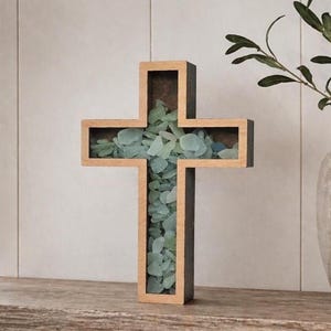May include: A wooden cross-shaped decorative piece filled with sea glass. The cross has a light brown frame with a dark brown interior and is filled with various shades of blue-green sea glass. It sits on a wooden shelf.