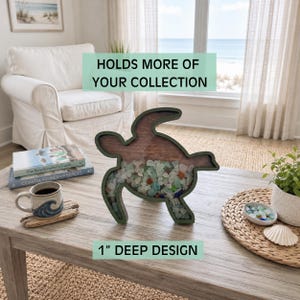 May include: A sea turtle-shaped shadow box with a dark green frame and a clear front panel, filled with assorted colorful sea glass. The text on the image reads: HOLDS MORE OF YOUR COLLECTION and 1" DEEP DESIGN. The object is placed on a wooden table.