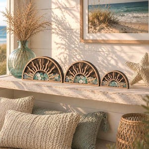 May include: Three semi-circular wooden sun-themed decorative displays of varying sizes, each featuring a cutout sunburst design filled with small, smooth, light blue and white sea glass pieces, arranged in a row on a white wooden surface.