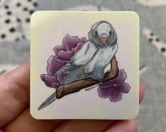 Puede incluir: Una pegatina con una ilustración dibujada a mano de un periquito azul y gris posado en una rama con flores moradas. El periquito tiene un cuerpo azul claro y una cabeza gris con un pico negro. Las flores son de un morado intenso y tienen un centro blanco.
