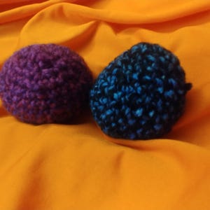 K&ouml;nnte beinhalten: Zwei geh&auml;kelte Kugeln, eine lila und eine blaue mit schwarzen Flecken, auf einer leuchtend orangenen Oberfl&auml;che.