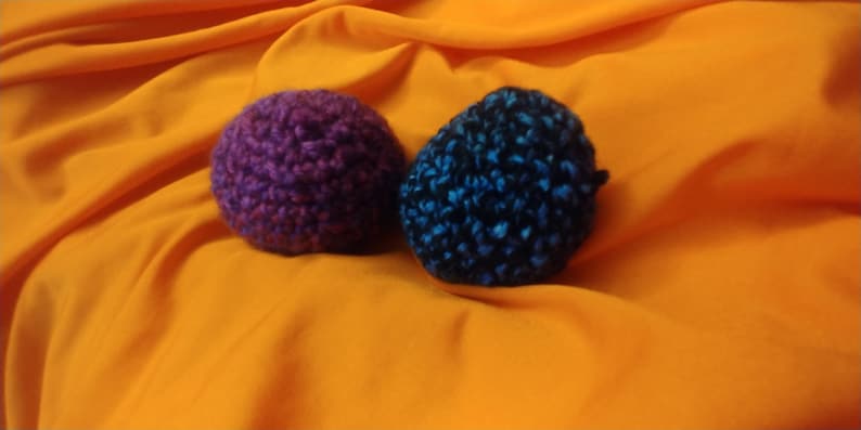 K&ouml;nnte beinhalten: Zwei geh&auml;kelte Kugeln, eine lila und eine blaue mit schwarzen Flecken, auf einer leuchtend orangenen Oberfl&auml;che.