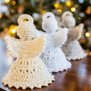 May include: Three crocheted angel ornaments with gold accents, displayed on a wooden surface in front of a blurred Christmas tree with lights.