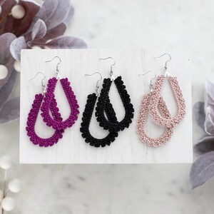 May include: Three pairs of crocheted dangle earrings in a white display stand. The earrings are in shades of pink, black, and purple.