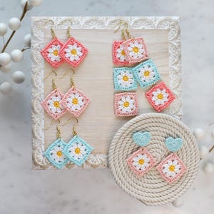 May include: A set of crocheted flower earrings in pink and blue. The earrings are displayed on a wooden board with a rope-like texture. The earrings are made with a delicate crochet pattern and feature a small white flower in the center.