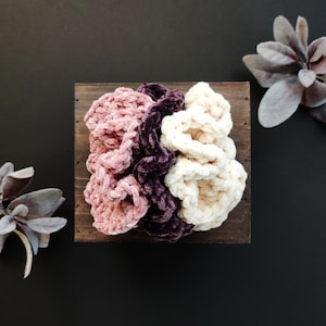 May include: Three crocheted hair scrunchies in a wooden box. The scrunchies are pink, purple, and white.