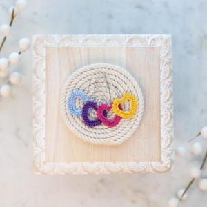 May include: Three crocheted heart-shaped earrings in blue, purple, and yellow on a white rope coaster. The earrings are hanging from silver hooks.